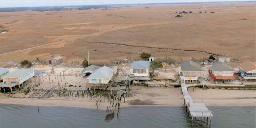 Aerial view of a coastal area with wooden houses on stilts, a sandy beach, and marshland in the background.