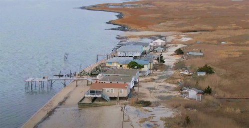 Aerial view of a coastal area with houses, a pier, and marshland along the shoreline.