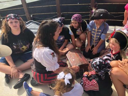 A group of children dressed as pirates gather around a treasure chest, listening to a pirate-themed storyteller.
