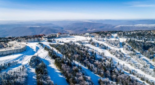 Aerial view of a snowy landscape with mountains, trees, and a resort nestled among the hills under a clear blue sky.