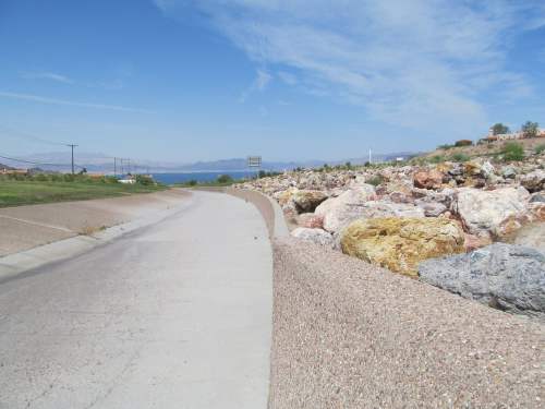 A wide, empty pathway alongside a rocky embankment, with a clear blue sky and distant mountains in the background.