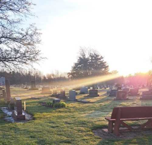 A serene cemetery at sunrise, with gravestones and a bench surrounded by trees and soft morning light.