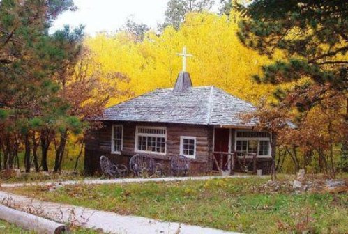 A rustic wooden cabin surrounded by vibrant autumn trees, featuring a cross on the roof and a pathway leading to it.