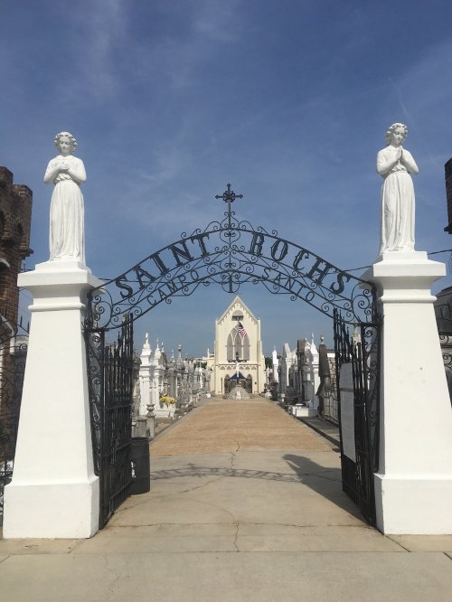 A wrought iron gate with "Saint Rochs" arching over a pathway leading to a church amidst a cemetery. Statues stand on either side.