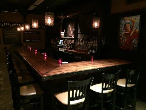 A dimly lit bar with a long wooden counter, high chairs, and decorative lanterns, featuring a colorful painting on the wall.