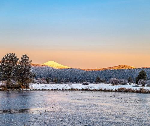 A serene winter landscape with snow-covered mountains, a frozen lake, and trees under a colorful sunrise sky.