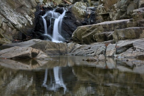 A serene waterfall cascades over rocks into a calm pool, reflecting the surrounding stone and nature.