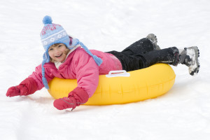 A smiling child in a pink jacket and hat sleds down a snowy hill on a yellow inner tube.