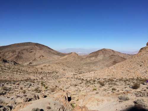 A vast desert landscape with rugged hills under a clear blue sky. Sparse vegetation is visible on the rocky terrain.