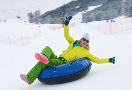 A person in a bright yellow jacket and green pants joyfully sleds down a snowy hill on a blue inner tube.