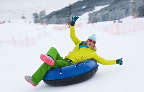 A person in a bright yellow jacket and blue beanie joyfully sleds down a snowy hill on a tube.