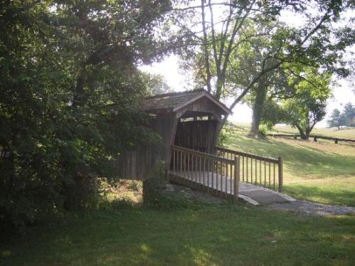 A rustic wooden covered bridge with a ramp, surrounded by trees and grassy fields in a serene landscape.
