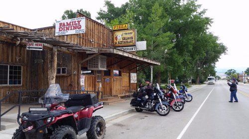 A rustic wooden restaurant with signs for dining and pizza, alongside parked motorcycles and an ATV on a quiet street.