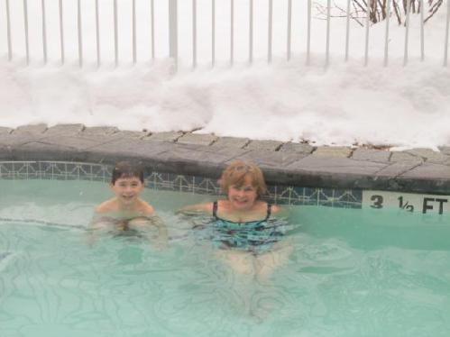 Two children play in a pool surrounded by snow, smiling and enjoying the water on a winter day.