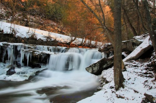 A serene waterfall surrounded by snow-covered rocks and trees, with warm light illuminating the scene.