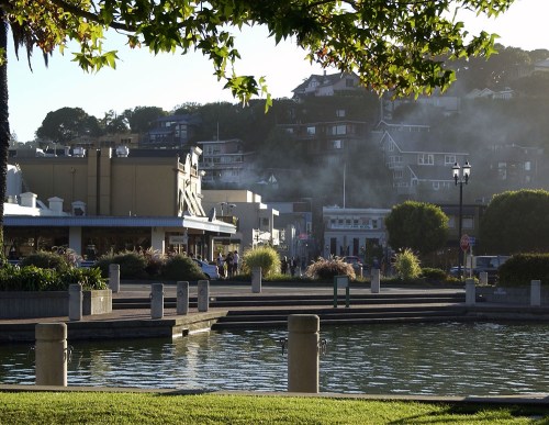A serene waterfront scene with buildings, greenery, and misty hills in the background, reflecting in the water.