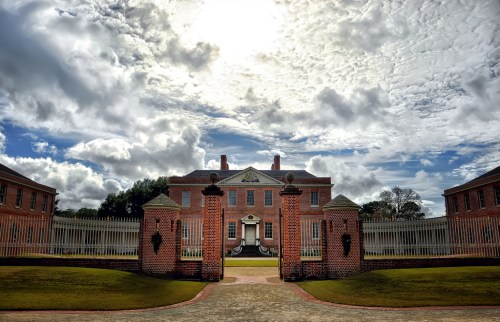 A grand brick mansion with a large gate, surrounded by lush green lawns and a dramatic cloudy sky.