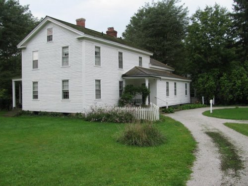 A white two-story house with a green lawn and a gravel driveway, surrounded by trees and shrubs.