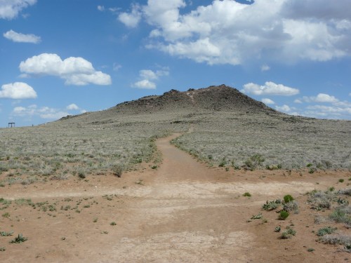 A dirt path leads through a grassy landscape towards a small hill under a blue sky with fluffy clouds.