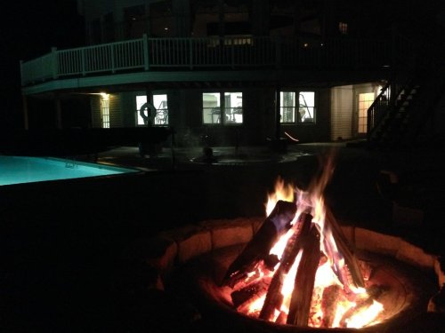 A cozy campfire in the foreground with a pool and a house illuminated at night in the background.