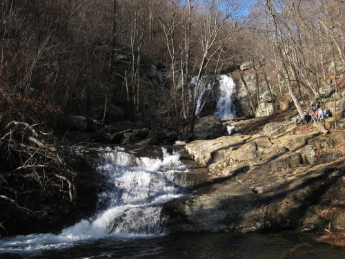 A serene waterfall cascades over rocks, surrounded by bare trees and a peaceful natural setting.