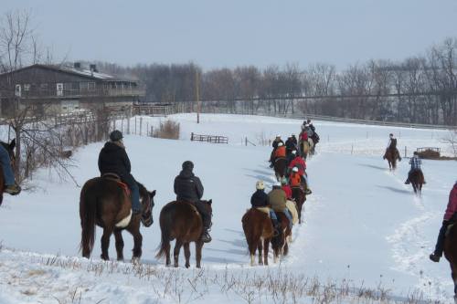 A group of people on horseback riding through a snowy landscape, with trees and a building in the background.