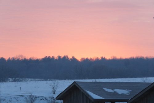 A serene winter landscape at dawn, featuring a colorful sky and snow-covered ground with silhouettes of trees in the distance.