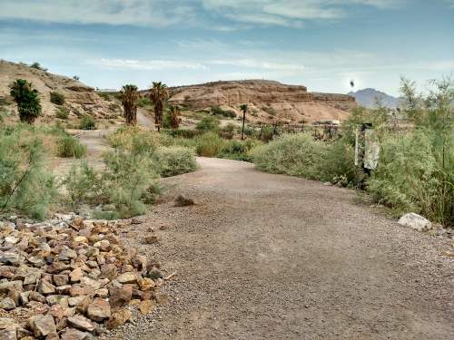 A winding dirt path surrounded by greenery and rocky terrain, with hills in the background under a cloudy sky.