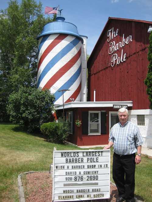 A man stands in front of a large barber pole building with a sign reading "World's Largest Barber Pole."