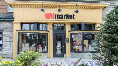 Front view of WV Market, featuring large windows, a yellow facade, and colorful flowers in the foreground.