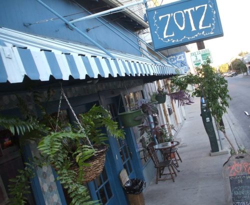 Exterior of Zotz café with hanging plants, blue walls, and a sign, featuring a sidewalk and street view.