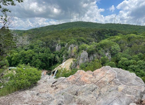 A scenic view of a lush green landscape with a waterfall, rocky foreground, and wind turbines on a distant hill.