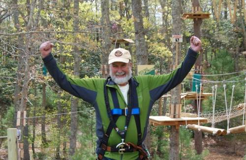 A smiling man in a harness raises his arms in celebration at an outdoor adventure park with trees and obstacles in the background.