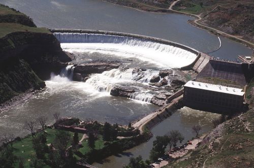 Aerial view of a large waterfall cascading over a dam, surrounded by green land and a river.
