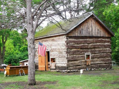 A rustic log cabin with a wooden door, an American flag, and surrounded by greenery and trees.