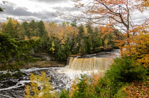 A scenic waterfall surrounded by autumn foliage and trees under a cloudy sky.