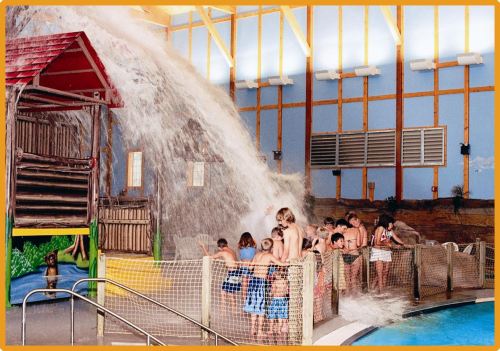 Children play in a water park with a large water feature, surrounded by colorful structures and safety fencing.