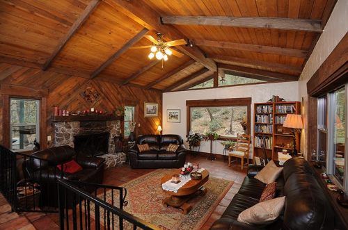 Cozy living room with wooden beams, stone fireplace, leather sofas, and a view of nature through large windows.