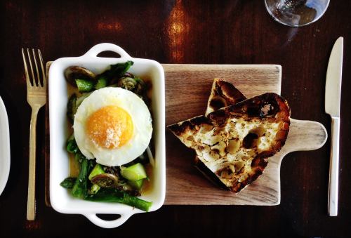A dish featuring a poached egg on vegetables, served with toasted bread on a wooden board. Fork and knife beside it.