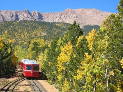 A red train travels along tracks surrounded by vibrant autumn trees and mountains in the background.