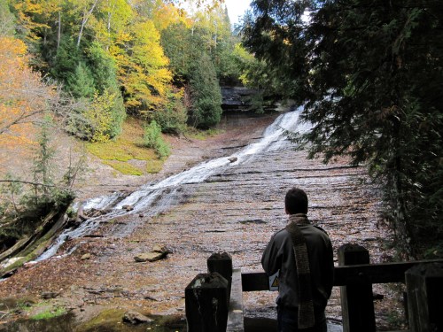 A person stands by a waterfall surrounded by autumn foliage, gazing at the cascading water.