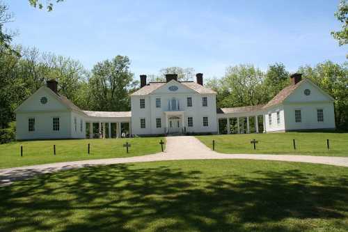 A large white historic house surrounded by green trees and a grassy lawn, with a clear blue sky above.