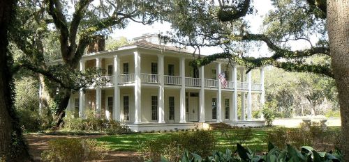 A large, white, two-story historic house with a porch, surrounded by trees and greenery. An American flag is displayed.