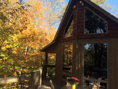 A wooden cabin with large windows surrounded by vibrant autumn foliage and a table with flowers on the deck.