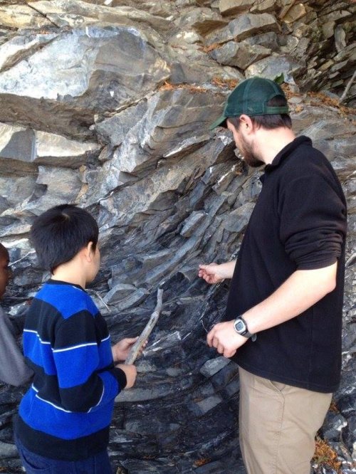 Two children and an adult examine rock formations, with one child holding a stick, in a natural outdoor setting.