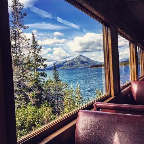 View from a train window showcasing a lake, mountains, and trees under a blue sky with fluffy clouds.