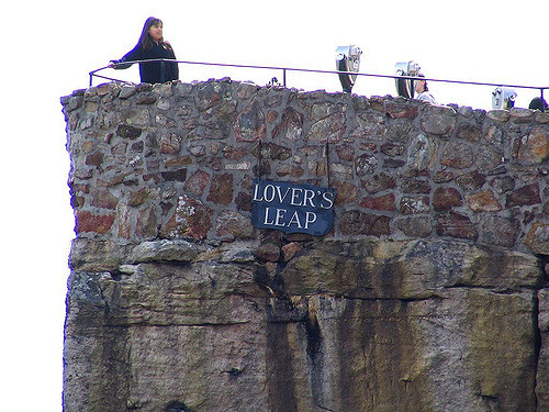A woman stands on a rocky cliff with a sign reading "Lover's Leap" above her, overlooking a scenic view.