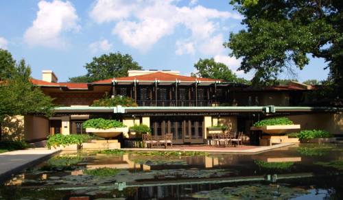 A modern building with a flat roof, surrounded by greenery and a pond, under a blue sky with fluffy clouds.