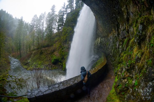 A person stands near a waterfall, surrounded by lush greenery and mist, with a river flowing below.
