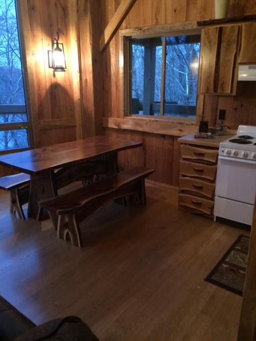 Cozy wooden kitchen with a dining table, benches, and a stove, featuring large windows and warm lighting.
